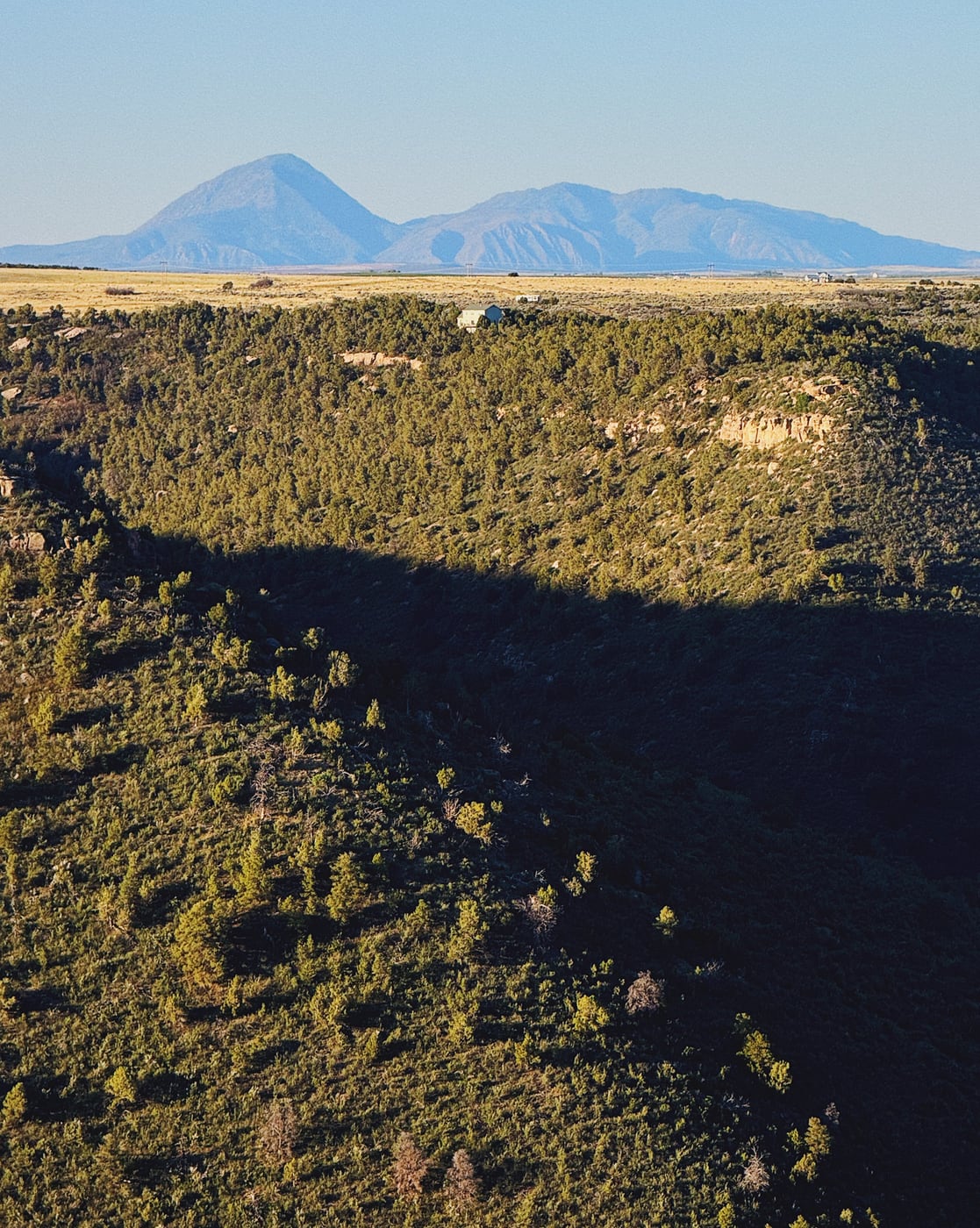 Distant mountain peaks with a grassland plateau and a forested, deep valley falling down to the bottom of the image in the foregrand. Under a clear, blue sky, a single, distant home is perched on the edge of the valley before it drops off.