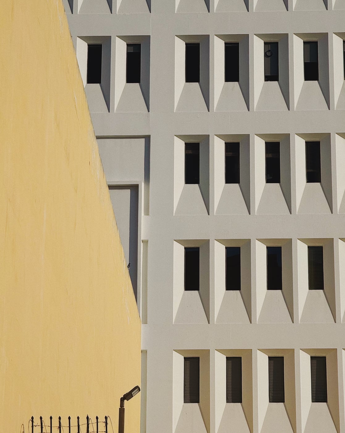 Facade of a modern building with repetitive geometric windows next to a yellow wall.