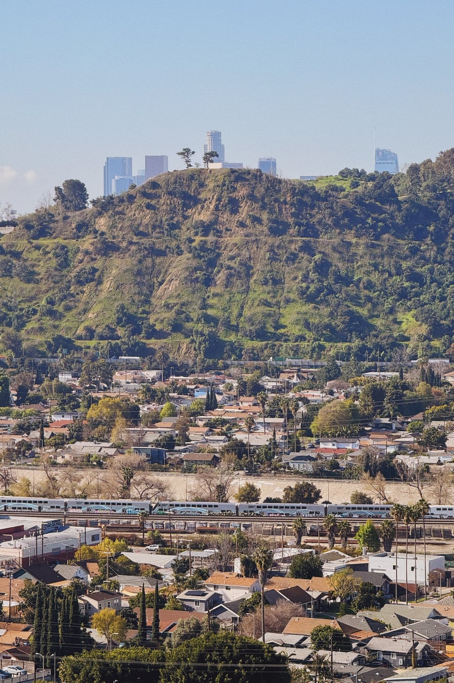 View of a hilly neighborhood with a train passing through in the foreground, and downtown Los Angeles city skyscrapers just visible in the distant background, peeking out above the hills.