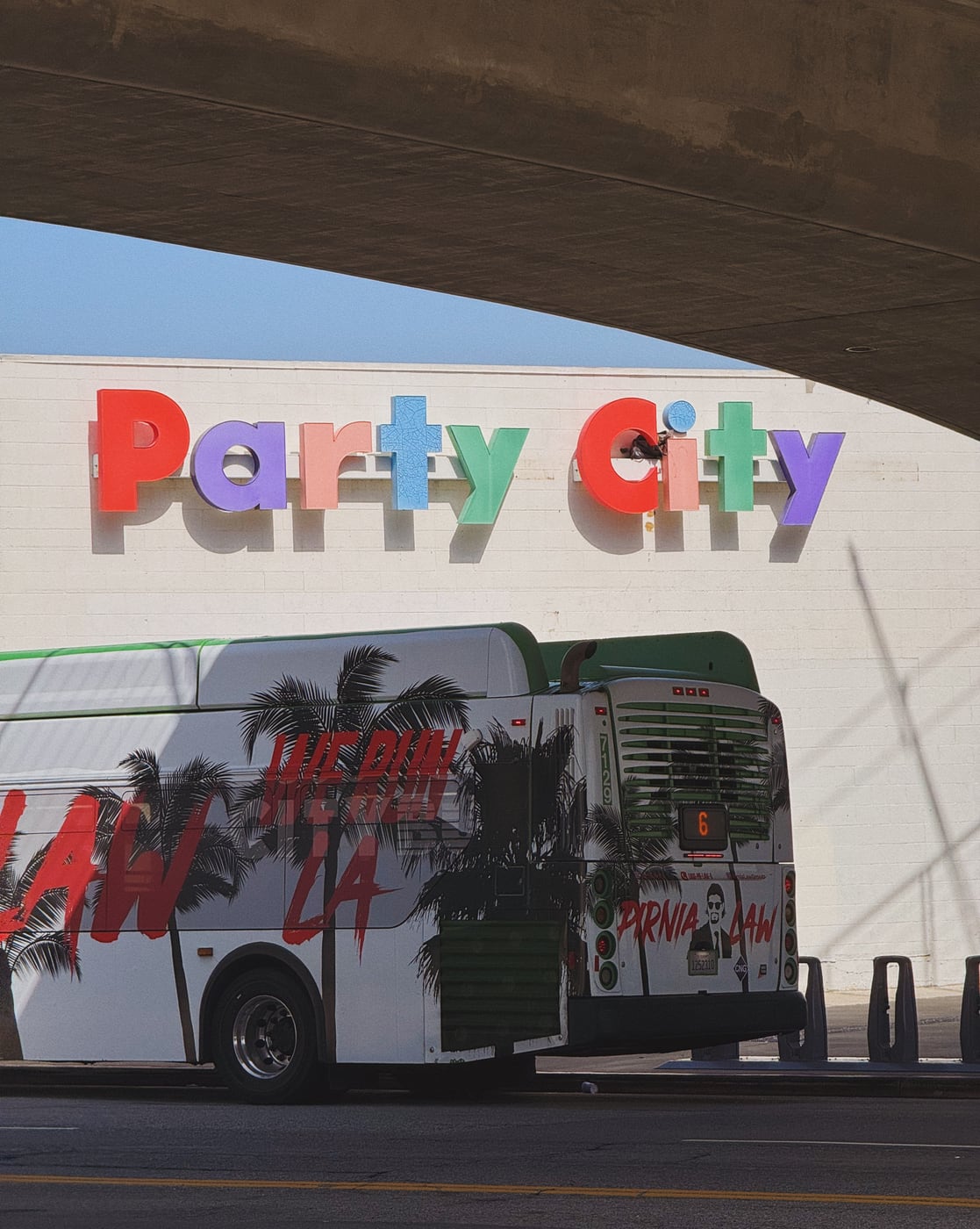 City bus with palm tree graphics parked in front of a Party City store sign under an overpass.