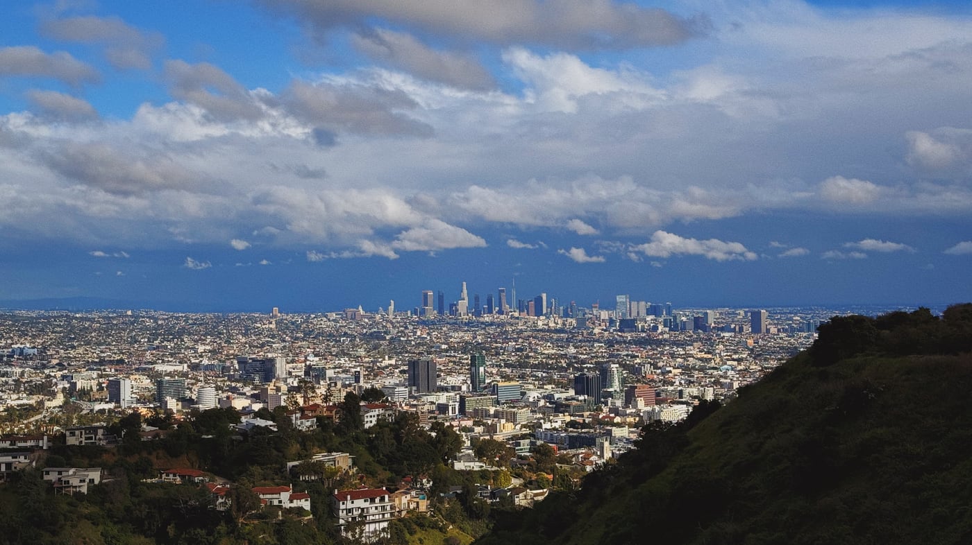 Panoramic view of the sprawling city of Los Angeles, with the downtown skyline in the distance, under a partly cloudy sky, with hills and residential areas in the foreground.