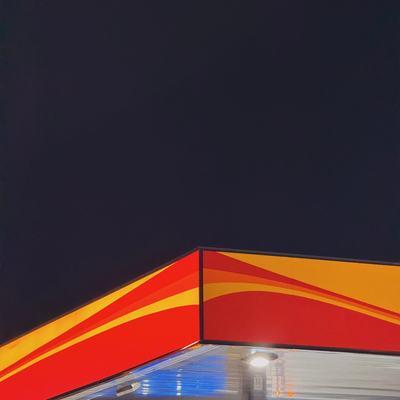 Brightly lit abstract corner of a gas station canopy with red and yellow stripes against a dark night sky.