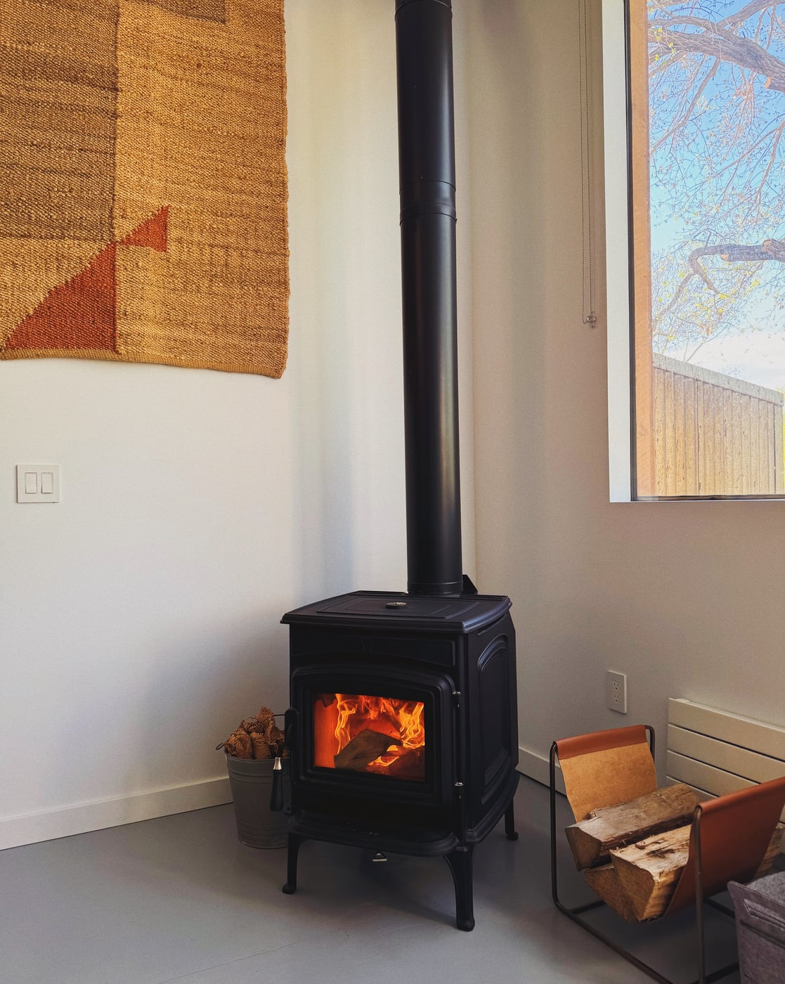 Cozy room with a wood-burning stove in the corner, stacked firewood to the right side, and a woven wall hanging to the left side. You see part of a window on the right side displaying a distant, winter tree and blue sky.