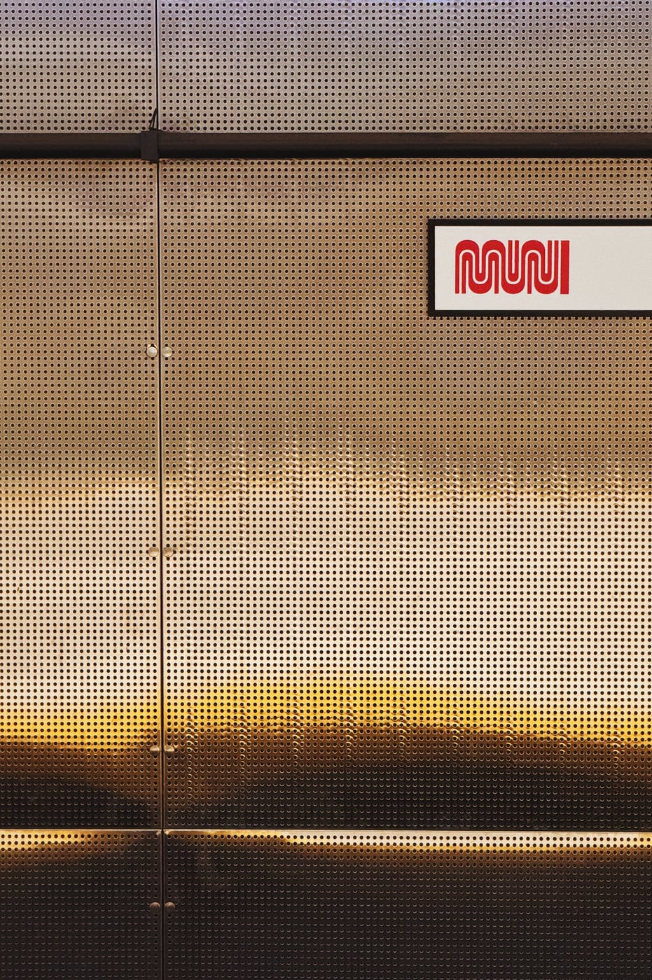 Perforated metal panel with a ‘MUNI’ logo in the upper right corner. The metal has a shiny gradient reflection moving between grey and gold.