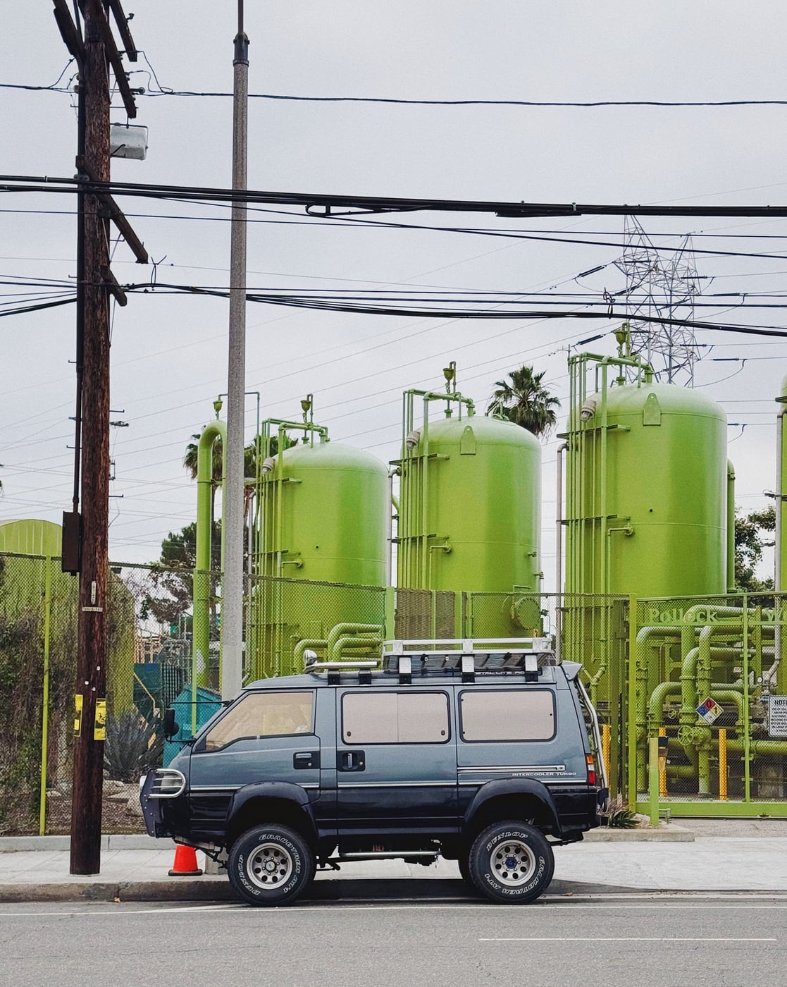 Van parked on a street in front of large green industrial tanks and pipes, with overhead power lines.