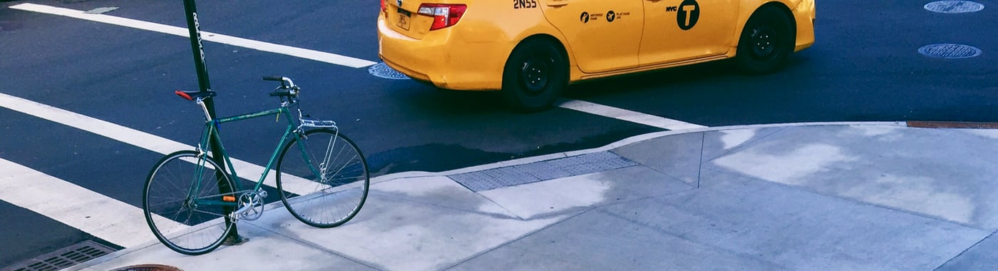 Concrete steps leading down to a corner sidewalk with a green bicycle parked on a street sign pole and an iconic NYC yellow taxi on the street.