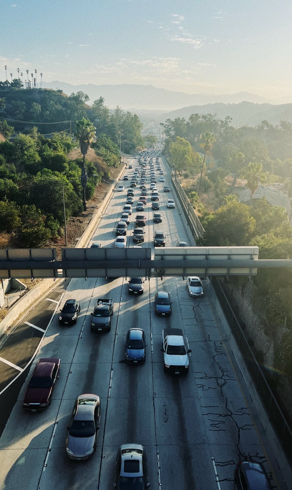 A top-down view of traffic on a multi-lane highway surrounded by green hills and trees, with mountains visible in the hazy distance.