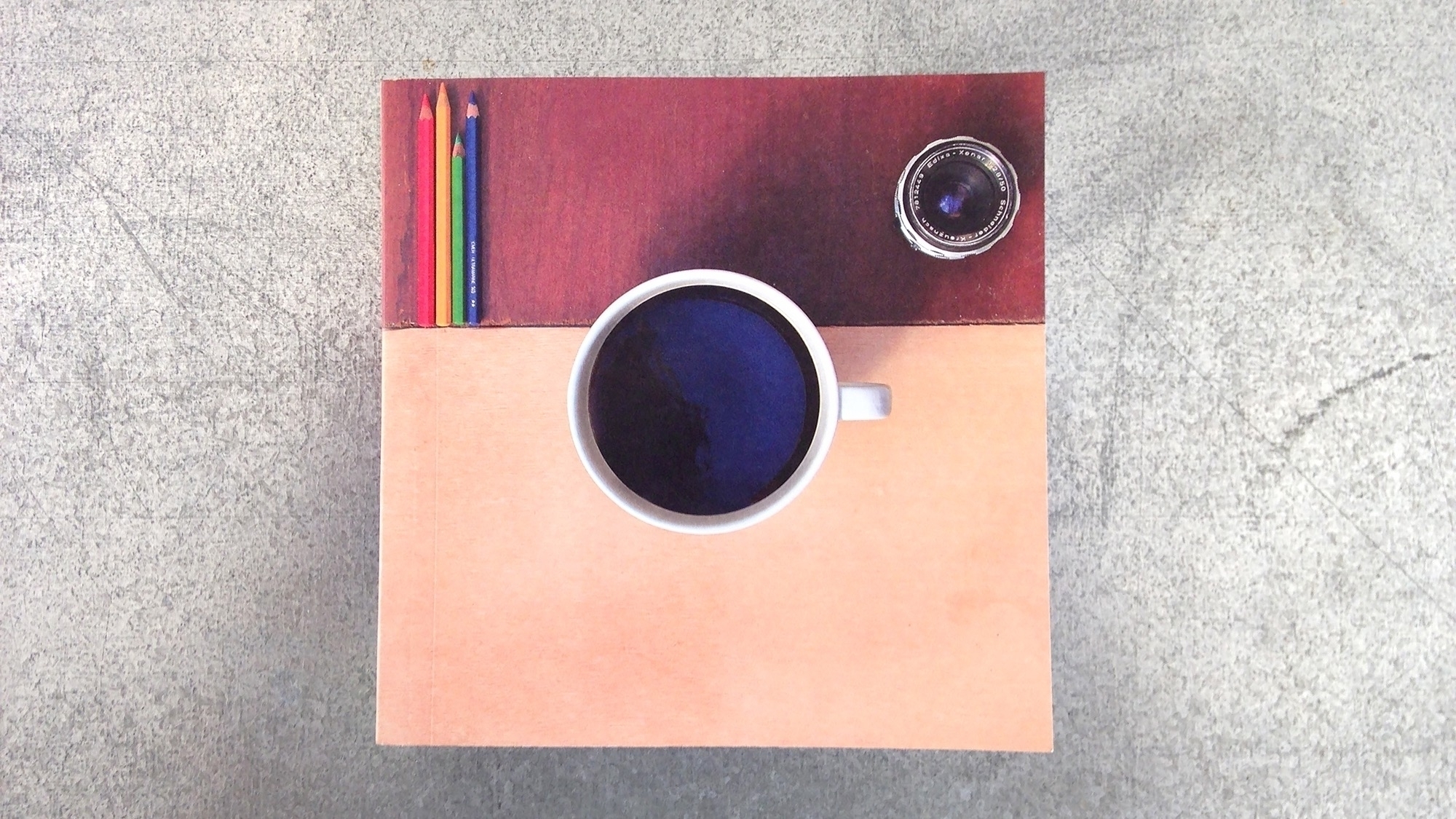 Top-down view of a white coffee cup filled with dark liquid, surrounded by colored pencils, a camera lens, and a two-tone background on a textured surface. Taken together, the objects look like the original Instagram app icon.